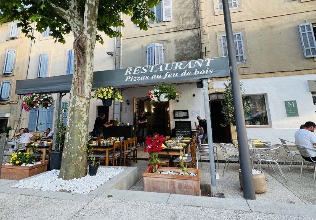 La façade du restaurant, enseigne « Pizzas au feu de bois », tables et chaises en terrasse sous les platanes